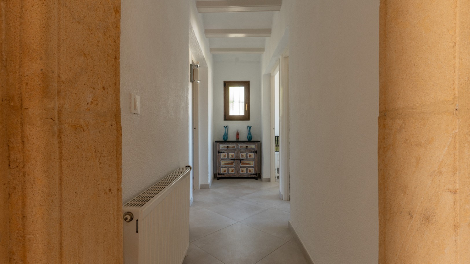 Pasillo luminoso con arcos de piedra tosca, vigas de madera blancas y suelos cerámicos. Incluye calefacción central y un aparador rústico bajo una ventana de madera.