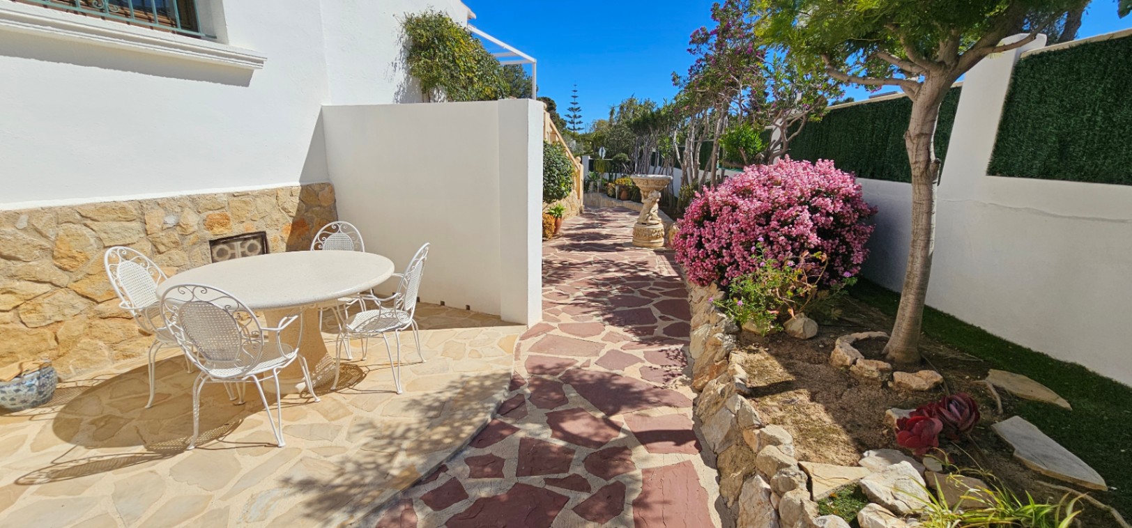 Patio exterior con pavimento de piedra natural, mobiliario de forja blanca y zona ajardinada con vegetación mediterránea y arbustos en flor.