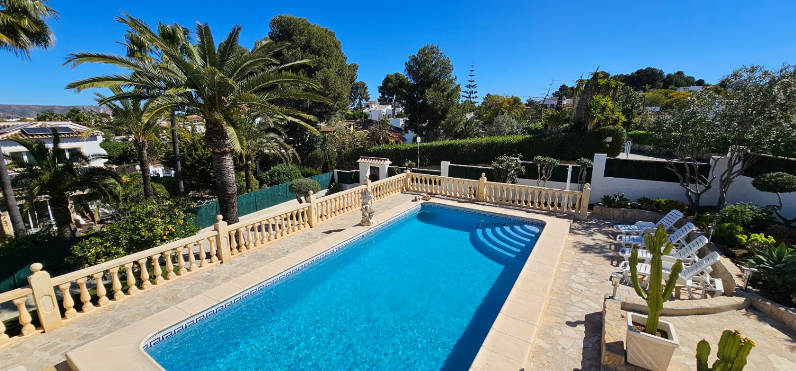 Piscina rectangular con escalera romana, rodeada de una terraza de piedra con balaustrada clásica y un jardín mediterráneo con palmeras maduras.