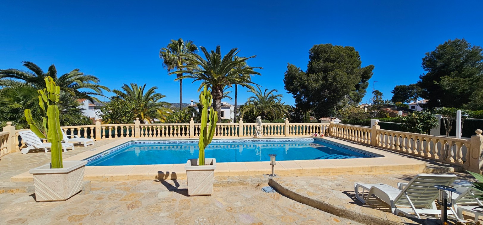 Piscina rectangular con escalera romana y cenefa decorativa, rodeada por una amplia terraza de piedra natural con balaustrada clásica y palmeras.