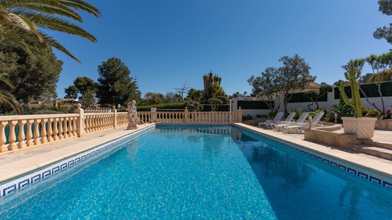 Piscina rectangular con revestimiento de gresite azul, rodeada por una terraza de piedra con balaustrada clásica y zona de hamacas entre palmeras y vegetación mediterránea.