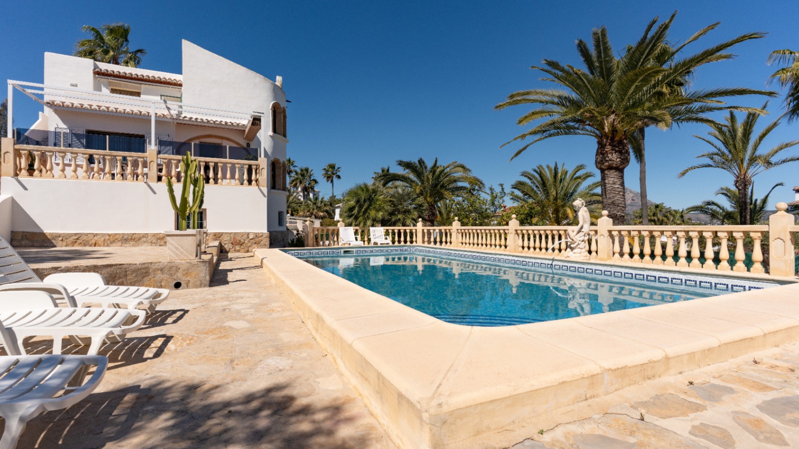 Piscina rectangular rodeada de una amplia terraza de piedra, balaustradas clásicas y palmeras, con la fachada blanca del chalet y sus terrazas al fondo.