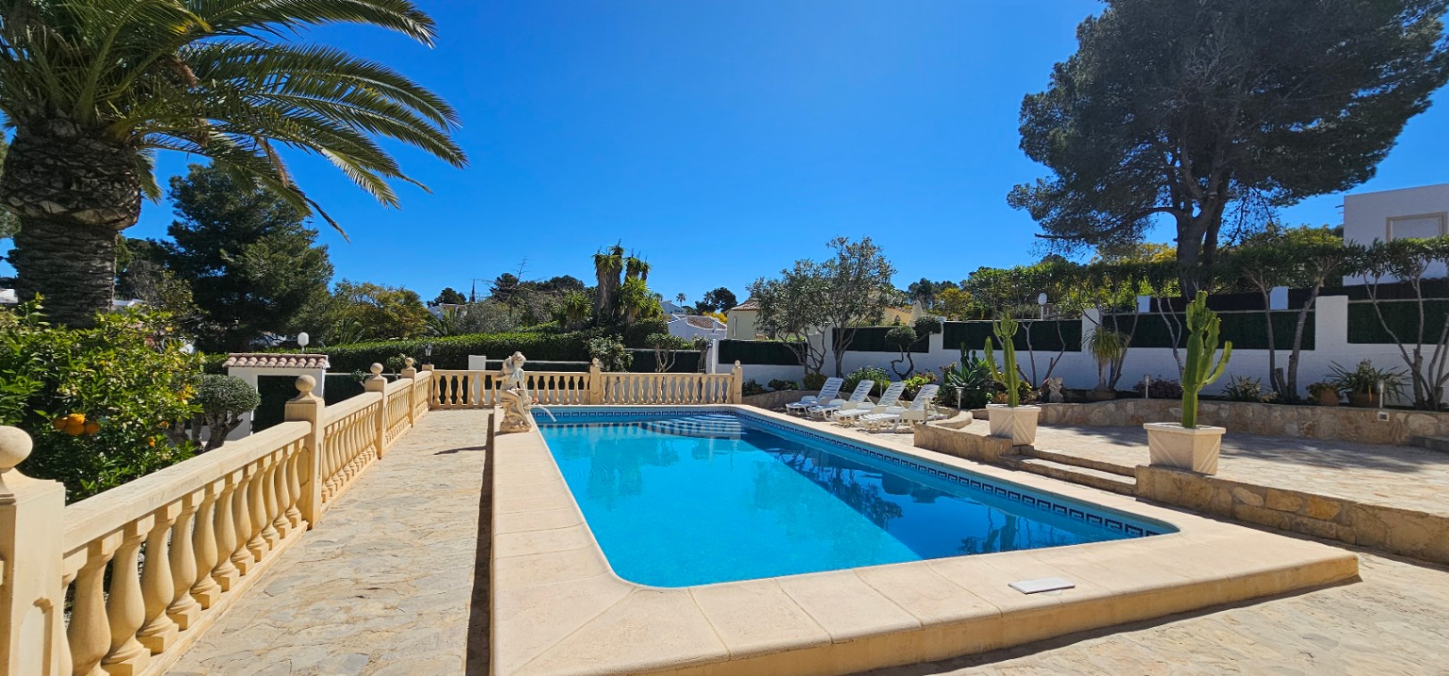 Piscina rectangular rodeada de una amplia terraza de piedra con balaustrada clásica, zona de tumbonas y vegetación mediterránea bajo un cielo despejado.
