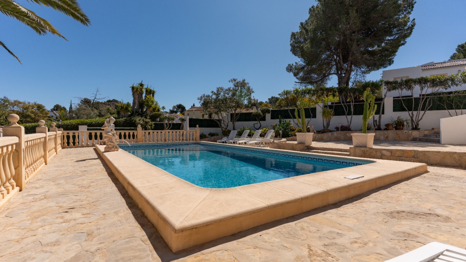 Piscina rectangular rodeada de una amplia terraza de piedra natural, balaustrada clásica y zona de solárium con tumbonas entre vegetación mediterránea.