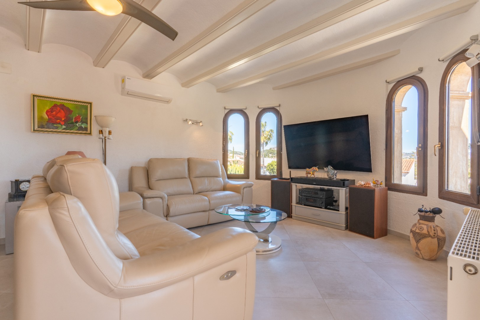 Salón con sofás de cuero crema, techo de vigas blancas y ventanas de arco. Cuenta con aire acondicionado, ventilador de techo y un completo sistema de entretenimiento.