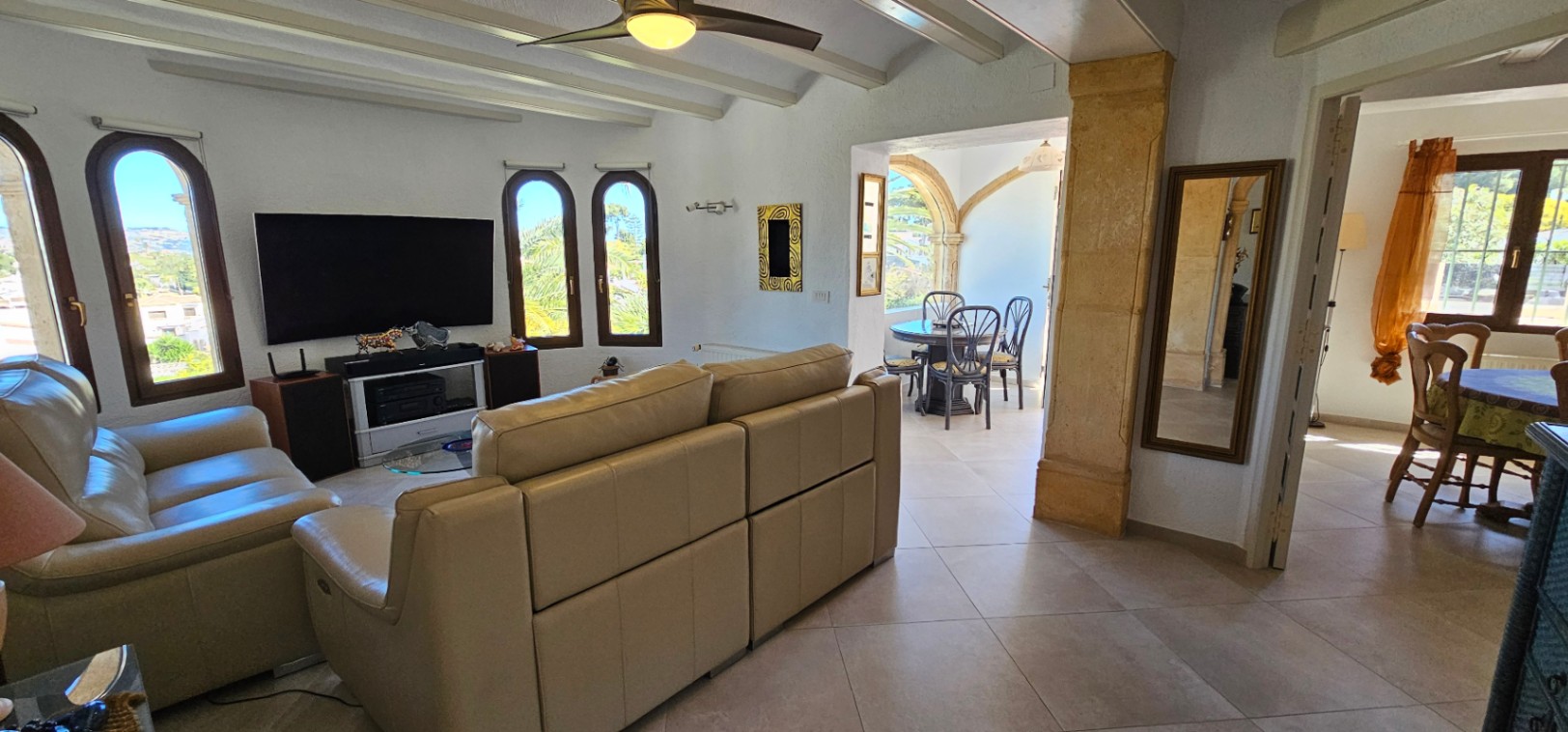 Salón con techos de vigas blancas, sofás de cuero beige y ventanas de arco. Un pilar de piedra tosca conecta el espacio con el comedor y la naya exterior.