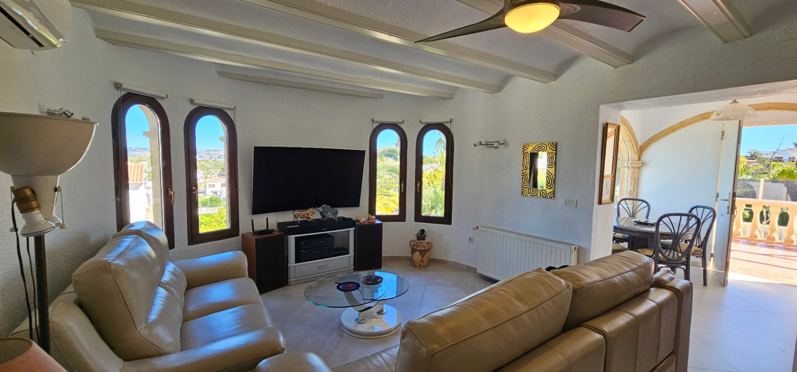 Salón con techos de vigas blancas, sofás de cuero y ventanales en arco. La estancia comunica mediante un arco de piedra con el comedor y la terraza exterior.