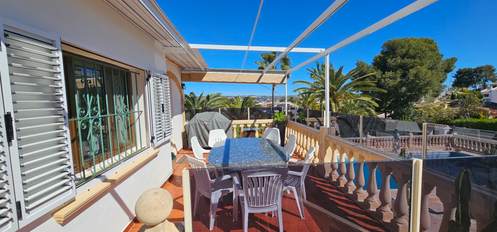 Terraza elevada con mesa de granito y toldo extensible, protegida por barandilla de cristal y piedra con vistas a la piscina y palmeras.
