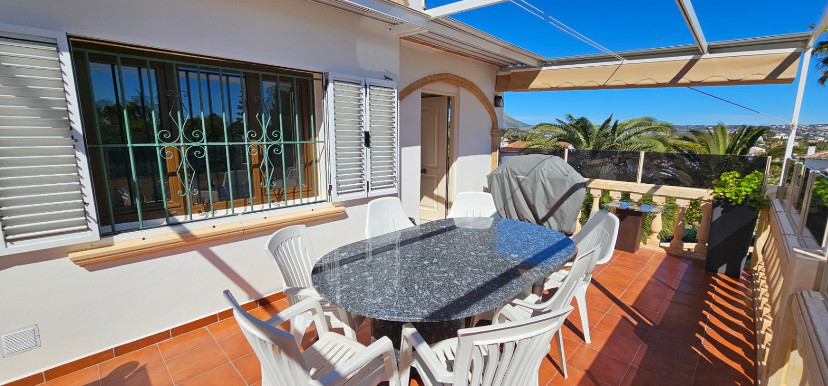 Terraza elevada con suelo de terracota, mesa de granito y vistas despejadas. Incluye toldo extensible, barandilla de piedra y acceso directo a la vivienda a través de un arco.