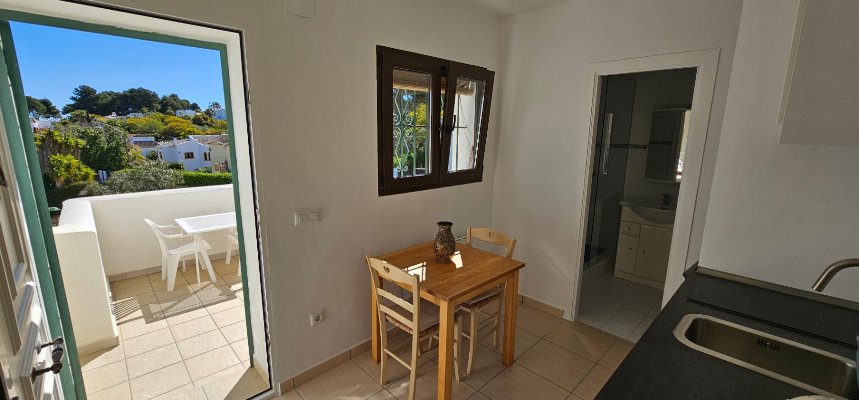 Zona de comedor con mesa de madera, acceso directo a la terraza soleada y vista parcial a la cocina y al baño privado.