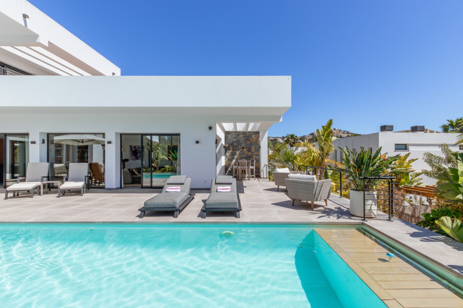Amplia terraza con piscina de gresite turquesa, zona de solárium y comedor exterior bajo pérgola, rodeada de vegetación mediterránea y arquitectura moderna blanca.