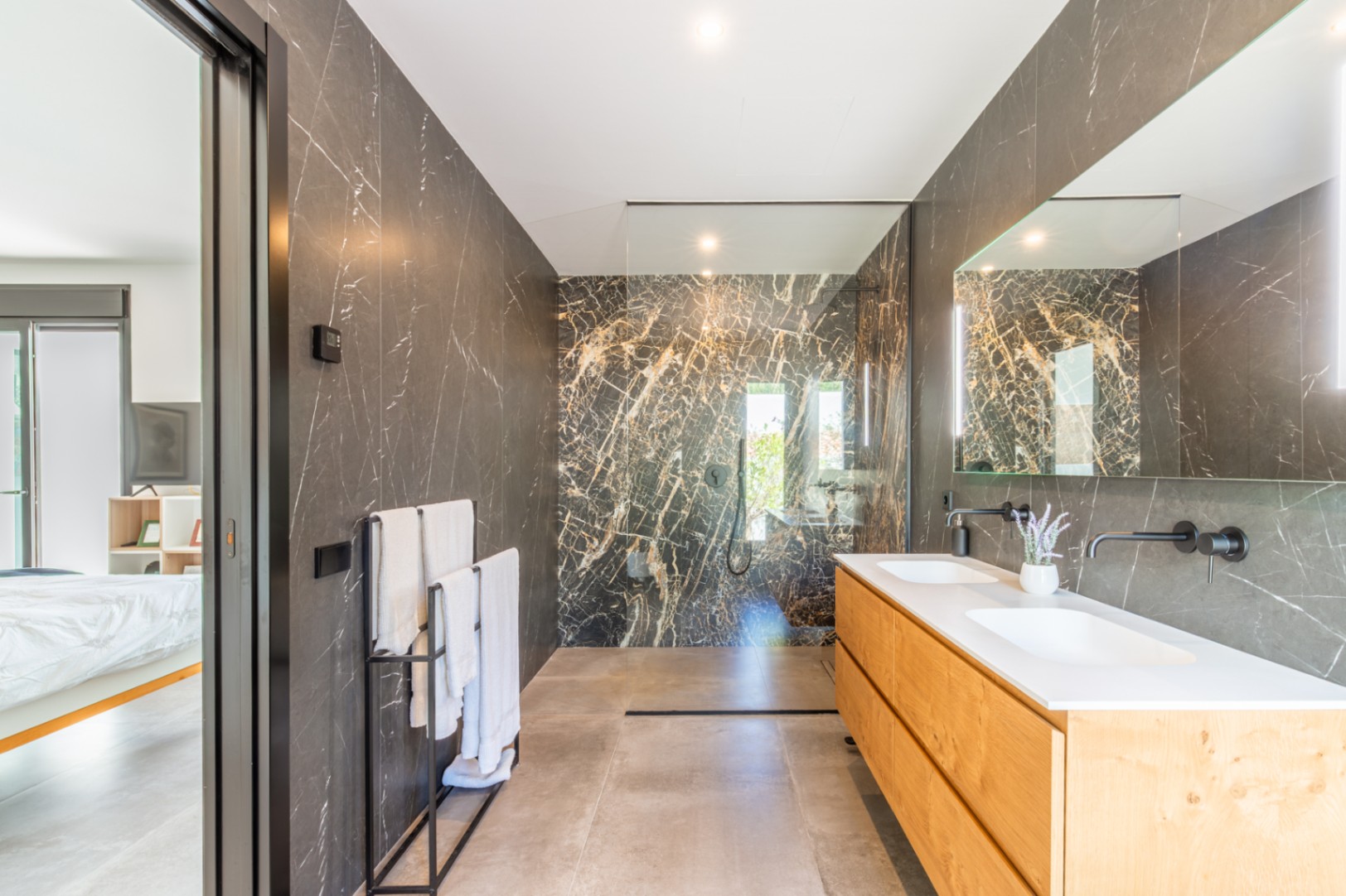 Baño principal con paredes de mármol oscuro, mueble de madera de doble seno y grifería negra empotrada, conectado directamente al dormitorio.