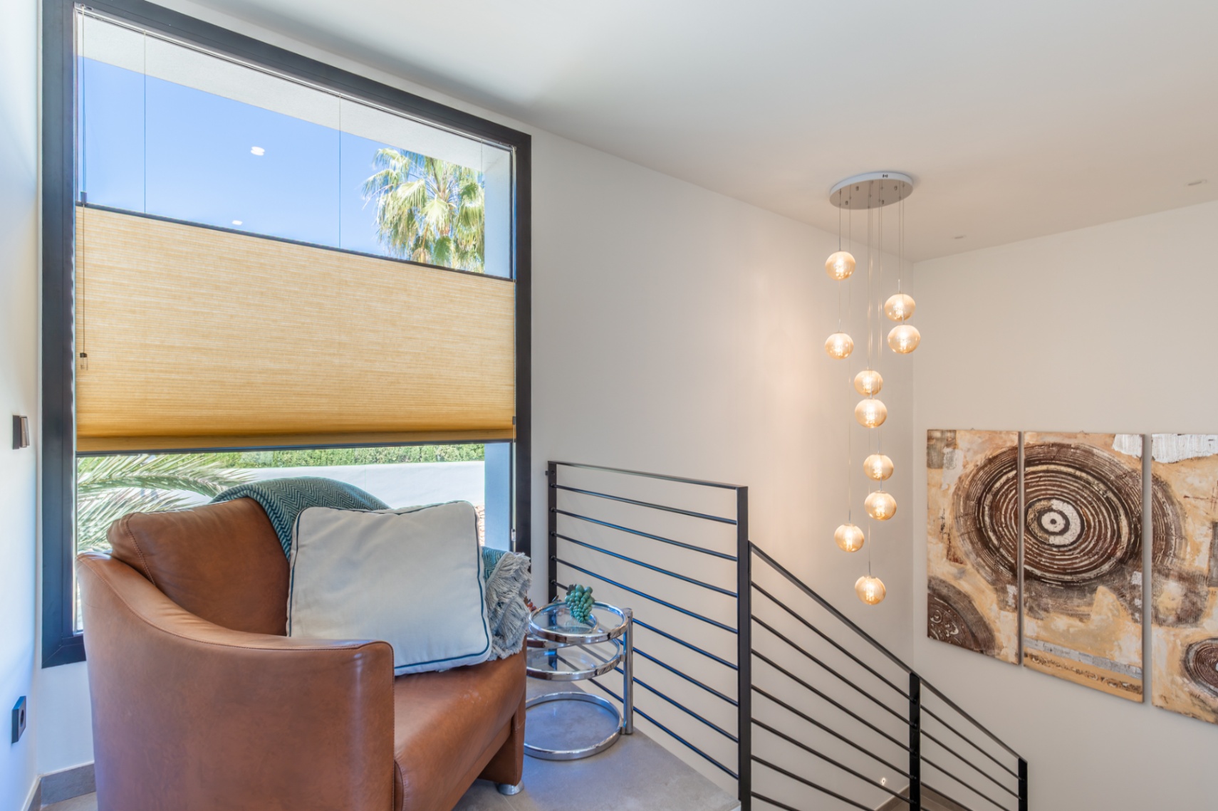 Rincón de lectura en el rellano superior con sillón de cuero, lámpara colgante de esferas y ventanal con persiana plisada y vistas a la vegetación exterior.