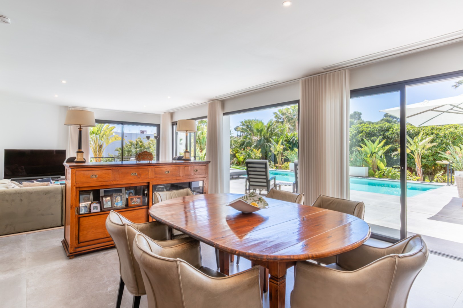 Salón-comedor diáfano con mesa de madera maciza y vistas directas a la piscina y al jardín mediterráneo a través de grandes ventanales.