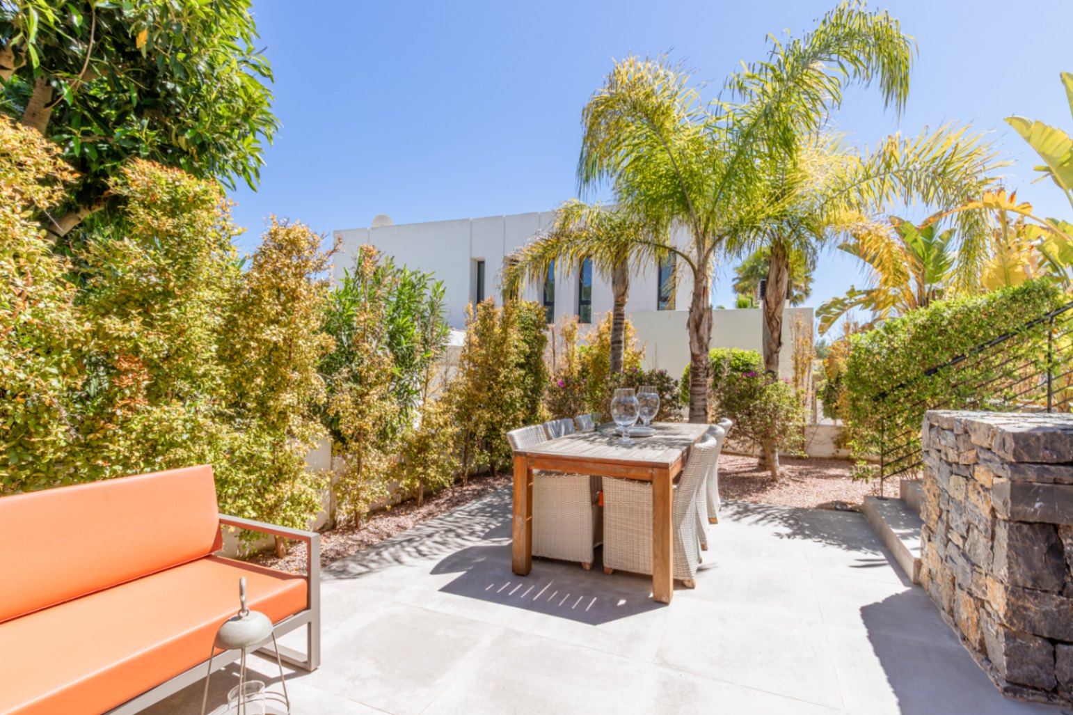 Terraza exterior con mesa de madera para seis comensales y sofá naranja, rodeada de vegetación madura y palmeras que garantizan privacidad.