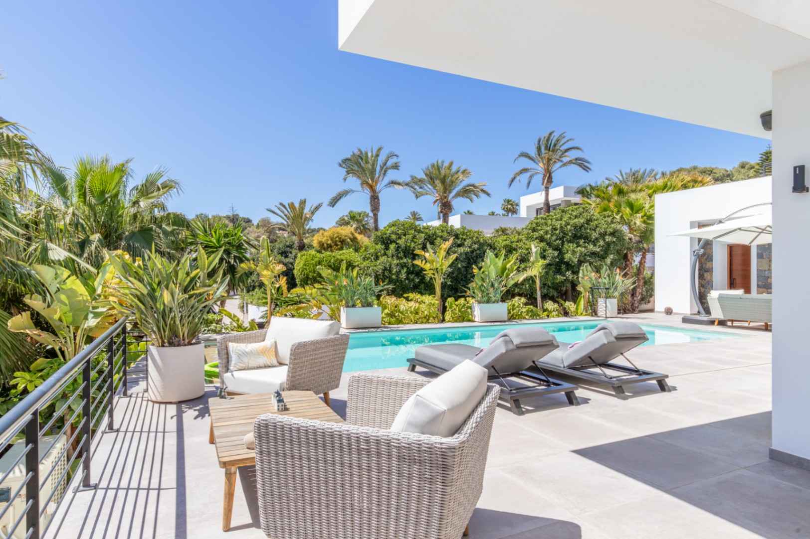 Terraza exterior con mobiliario de ratán y tumbonas frente a la piscina, rodeada de un jardín mediterráneo maduro con palmeras y vegetación privada.
