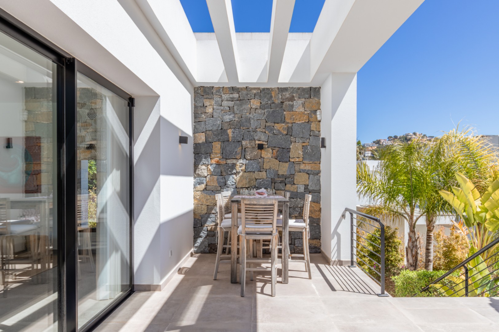 Terraza exterior con pérgola blanca, pared de piedra natural y zona de comedor elevada. Ofrece vistas despejadas y acceso directo al interior mediante grandes ventanales.
