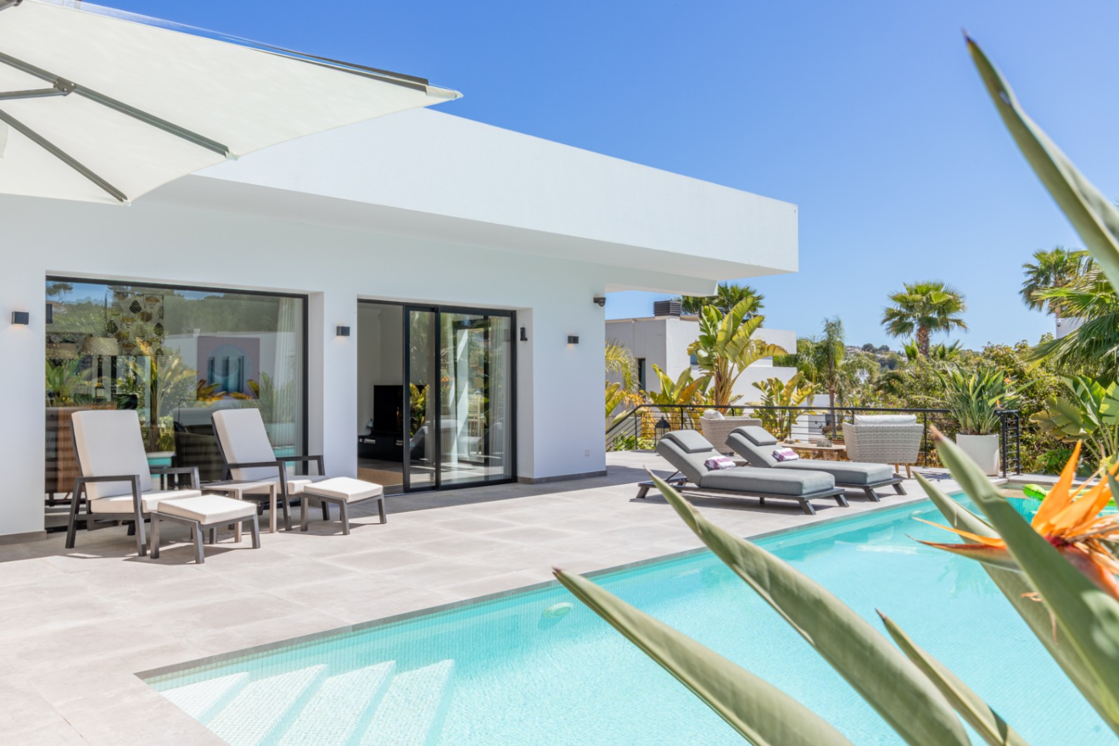 Terraza exterior con piscina de gresite claro, tumbonas de diseño y acceso directo al salón mediante ventanales de carpintería negra bajo una arquitectura blanca minimalista.