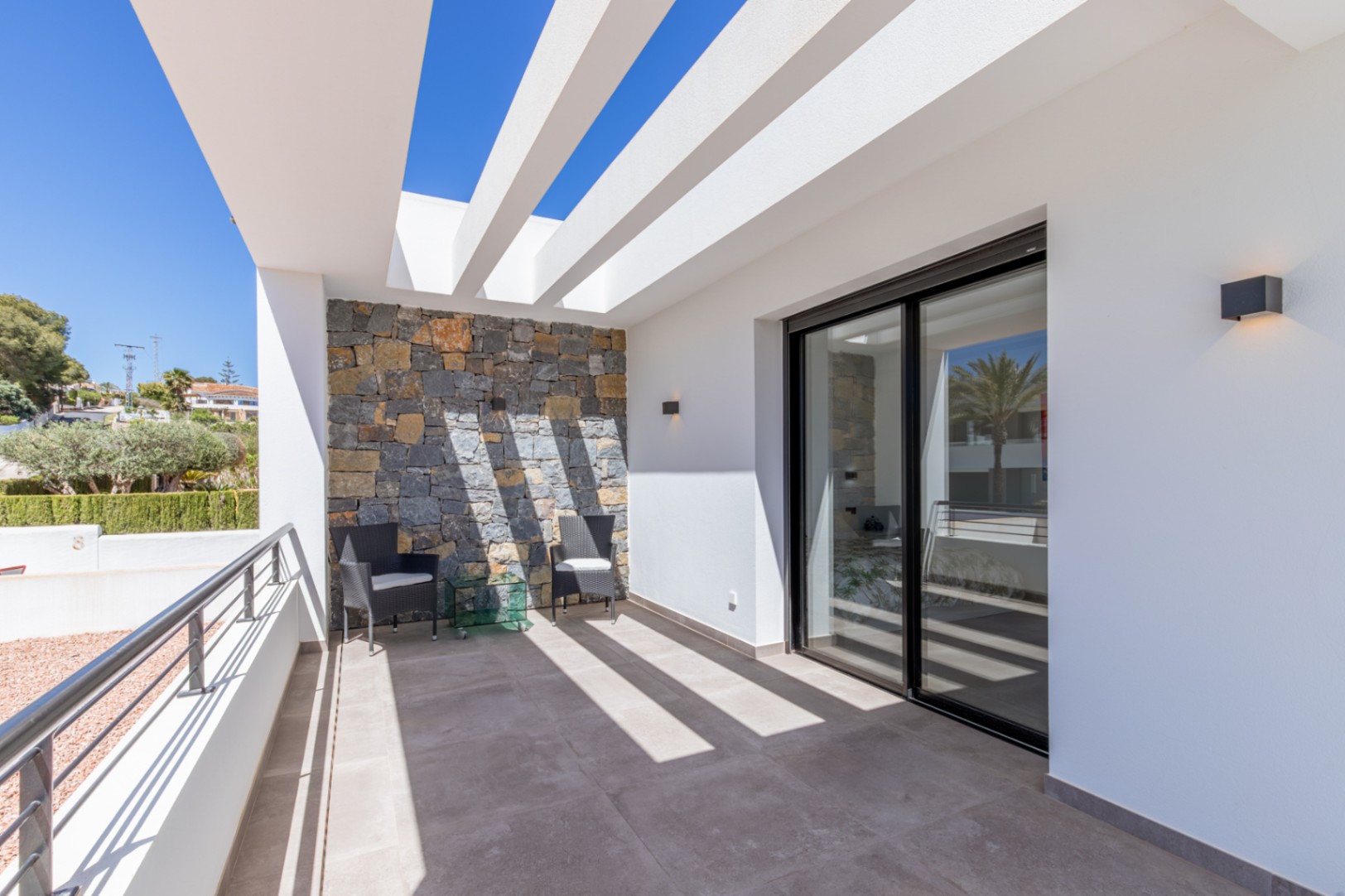 Terraza privada con pérgola de vigas blancas, pared de piedra natural y acceso directo a través de grandes ventanales correderos con marcos negros.