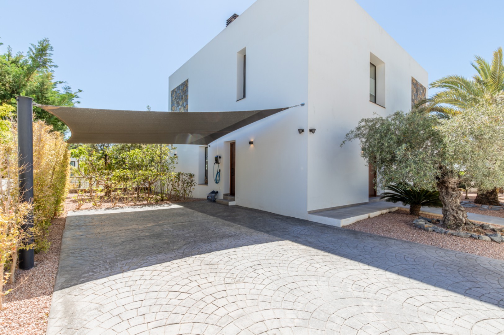 Zona de aparcamiento con pavimento impreso, vela de sombra y punto de carga para vehículos eléctricos junto a la entrada lateral de la villa.