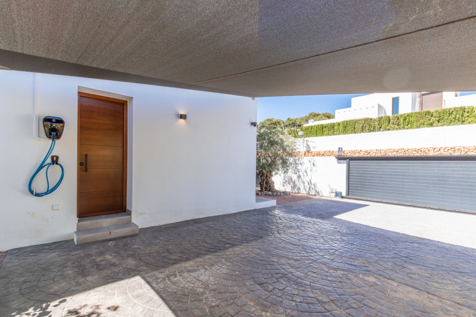 Zona de aparcamiento protegida por un toldo de vela, con suelo de hormigón impreso, punto de carga para vehículos eléctricos y acceso directo a la vivienda.