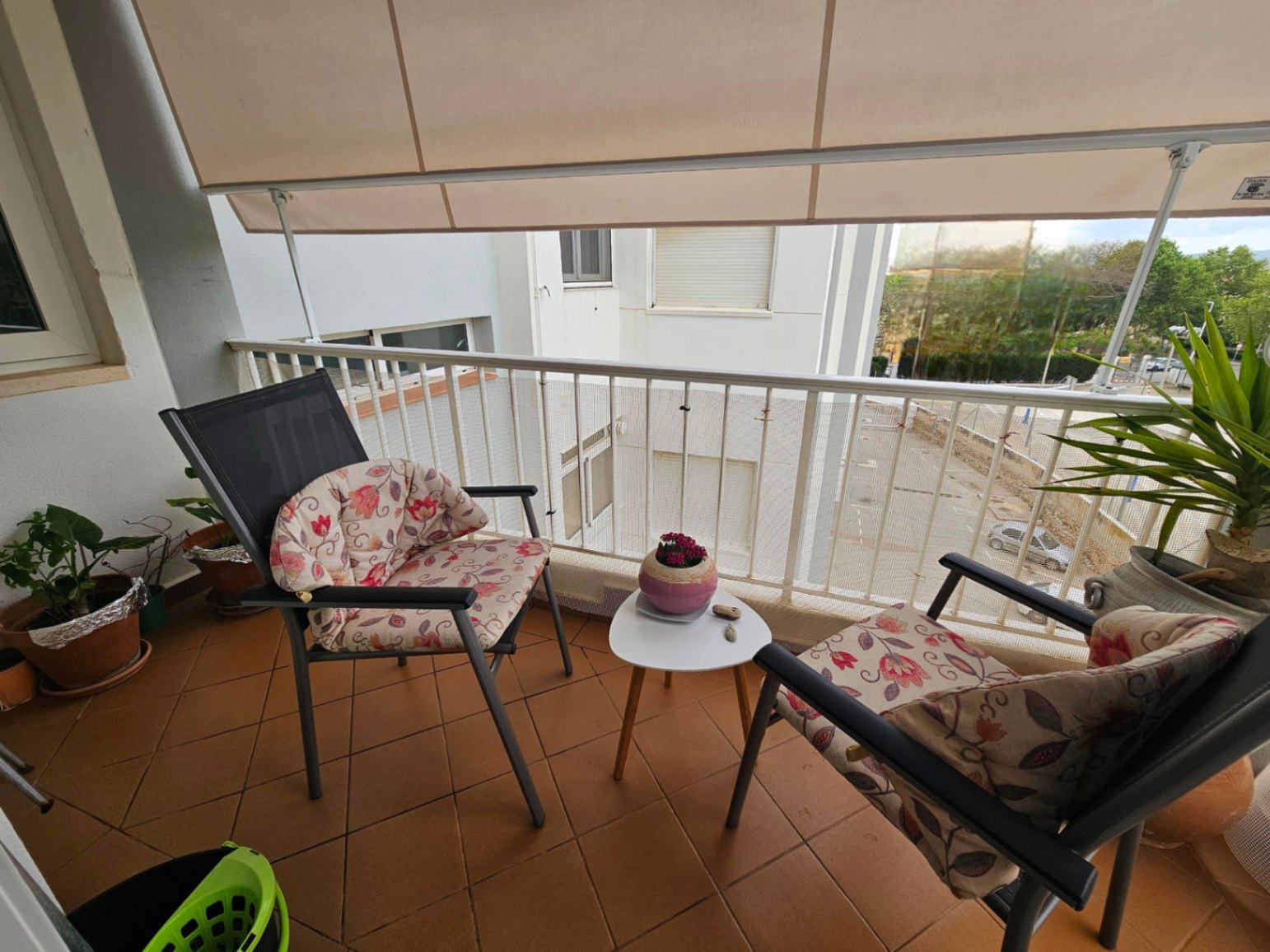 Balcón con suelo de baldosas de terracota, equipado con un toldo extensible y mobiliario de exterior compuesto por dos sillas con cojines y una mesa auxiliar.