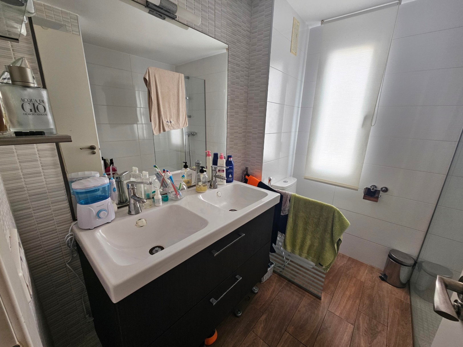 Baño moderno con mueble de doble lavabo en tono oscuro, gran espejo de pared y suelos de gres efecto madera. La estancia cuenta con luz natural y paredes alicatadas en tonos neutros.