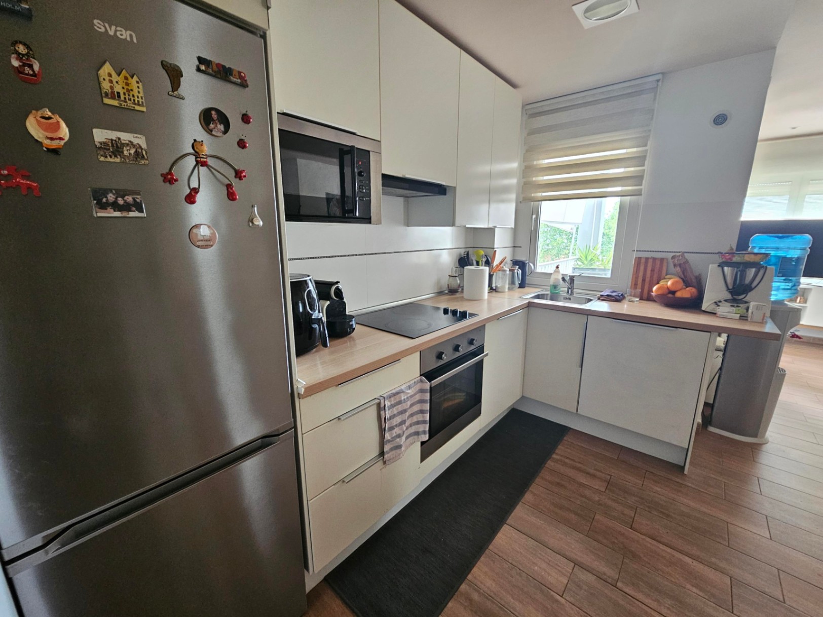 Cocina funcional en forma de L con encimera de madera, electrodomésticos integrados y ventana exterior. El suelo de gres cerámico imita la calidez de la madera.