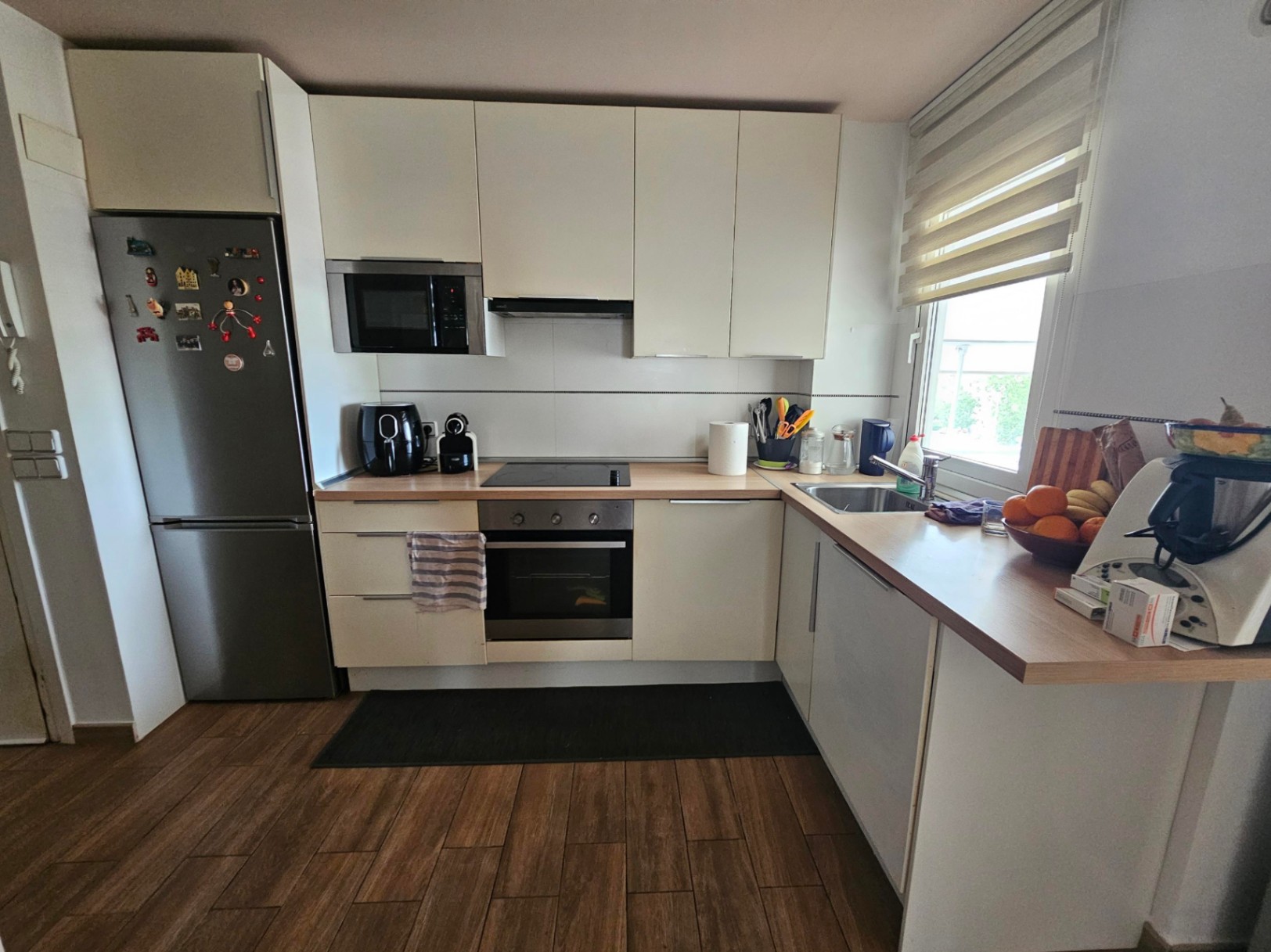 Cocina moderna en forma de L con armarios blancos, encimera de madera clara y electrodomésticos de acero inoxidable sobre suelo cerámico efecto madera.