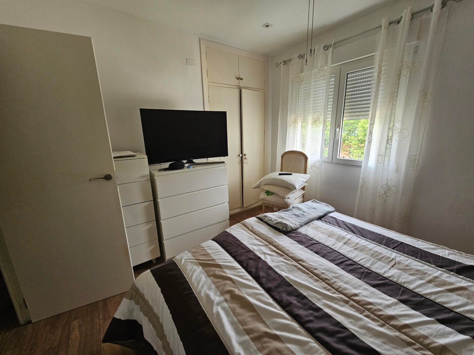 Dormitorio funcional con armarios empotrados, cómodas blancas y una ventana con persianas que permite la entrada de luz natural sobre el suelo laminado.