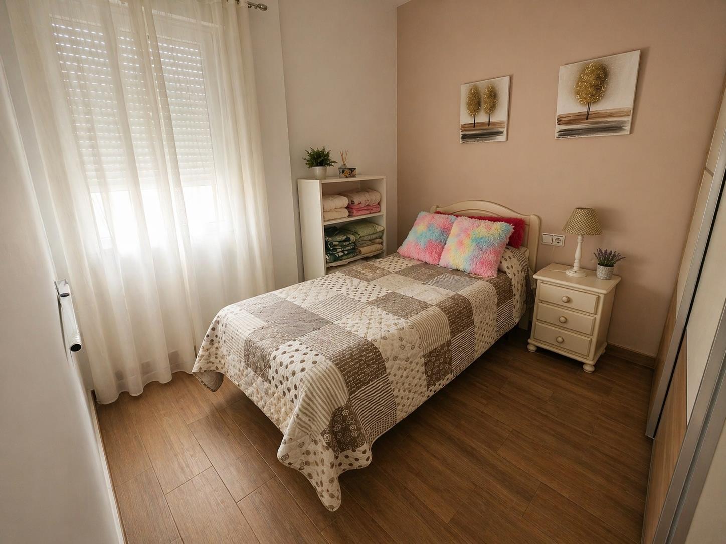 Dormitorio individual con suelos de gres efecto madera, ventana con persiana y cortinas blancas, y una pared de acento en tono tierra.