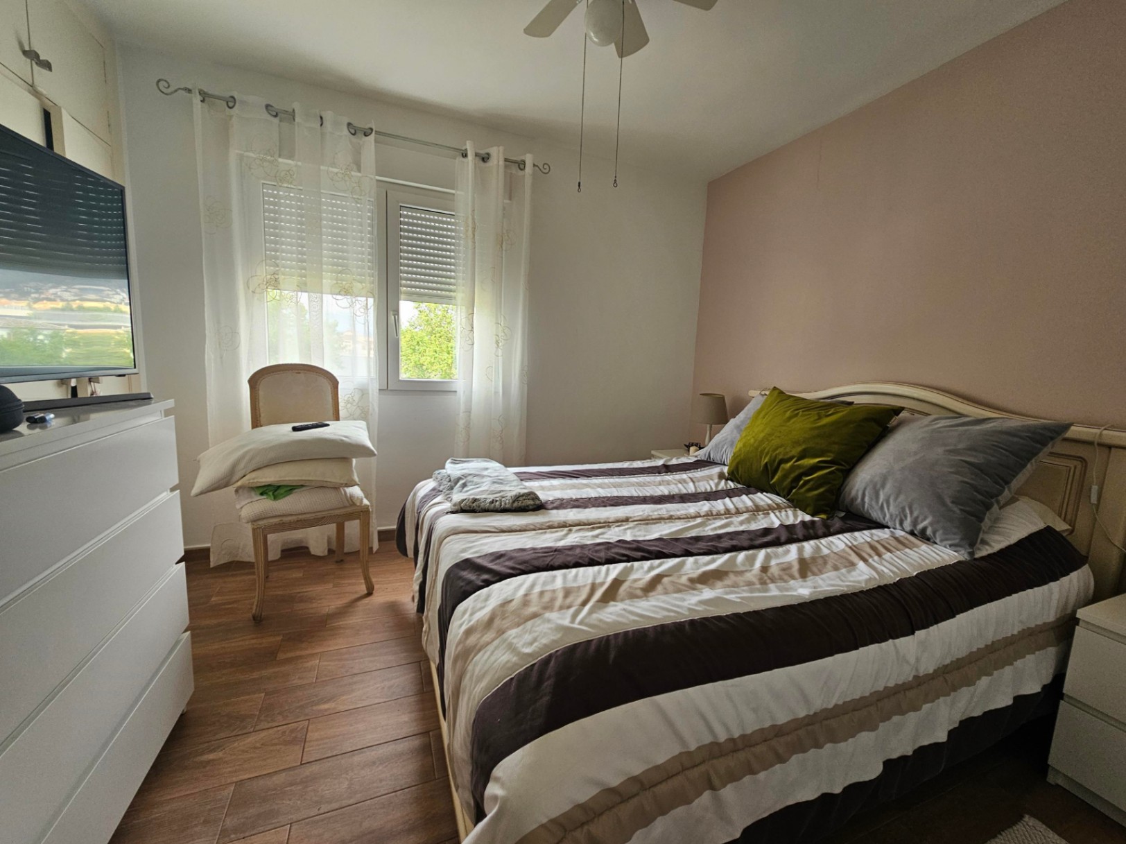 Dormitorio luminoso con suelos de gres efecto madera, pared de acento en tono salmón y ventana con persiana. Incluye ventilador de techo y mobiliario funcional blanco.