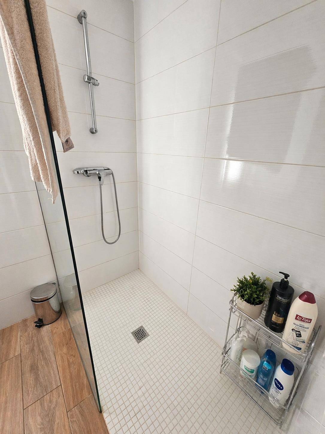 Ducha de estilo italiano con mampara de cristal, revestimiento de azulejos blancos horizontales y suelo de mosaico. Grifería cromada y suelo exterior efecto madera.