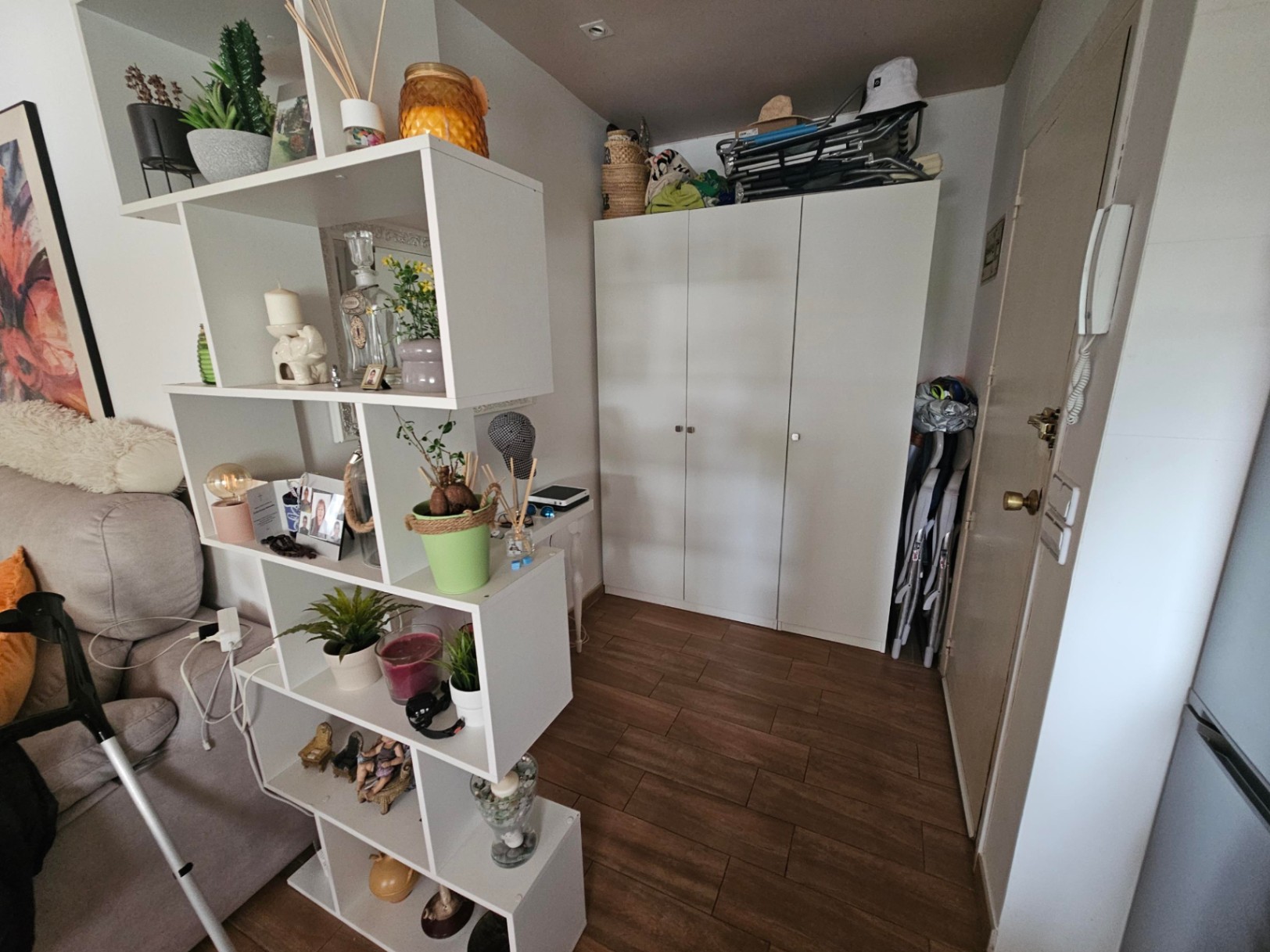 Hall de entrada funcional con suelos de gres efecto madera, armario de tres puertas blanco y estantería divisoria asimétrica con elementos decorativos.