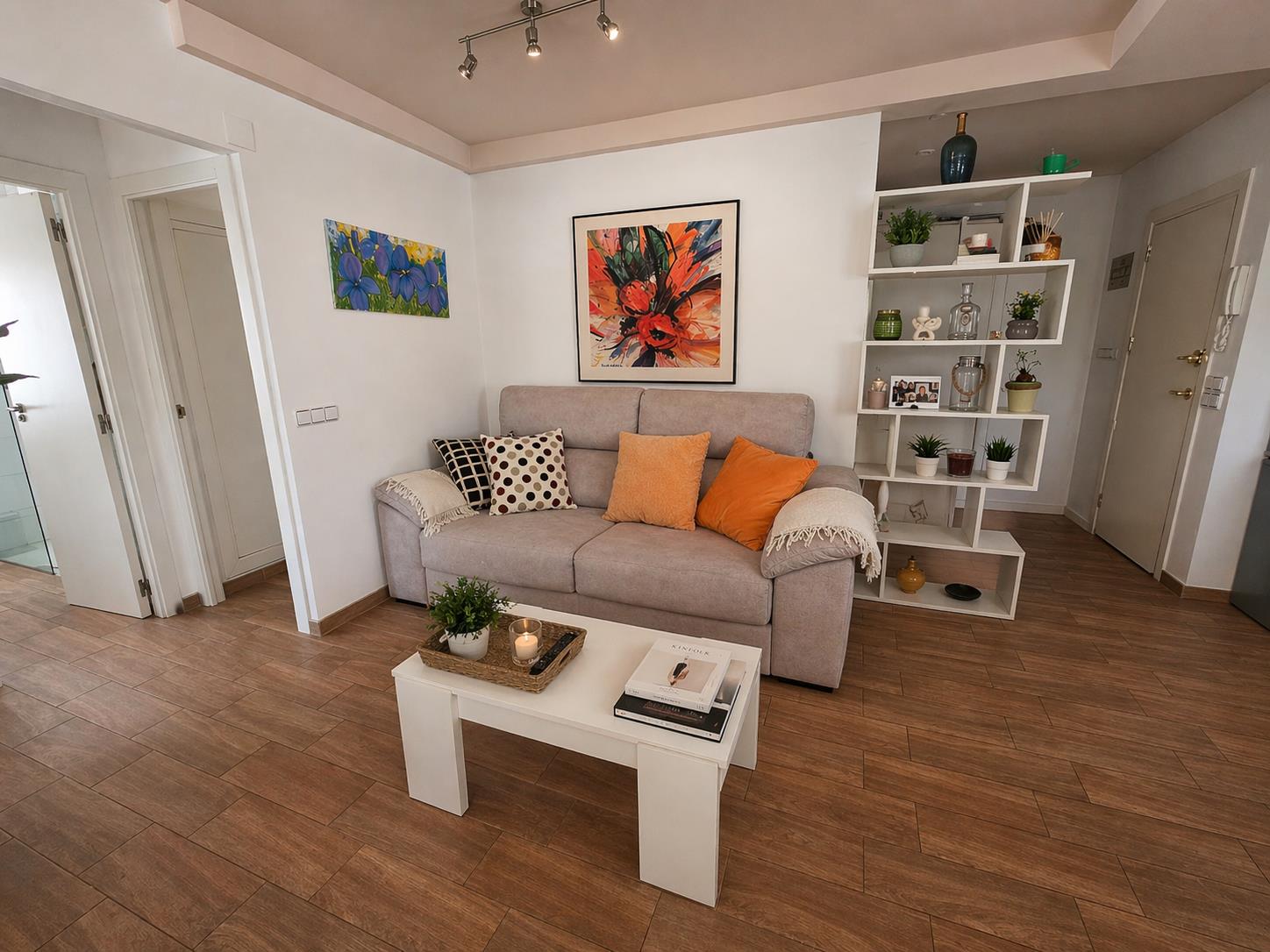 Salón acogedor con sofá de tela, mesa de centro blanca y suelos de gres efecto madera. Incluye una estantería geométrica decorativa y acceso a los dormitorios.