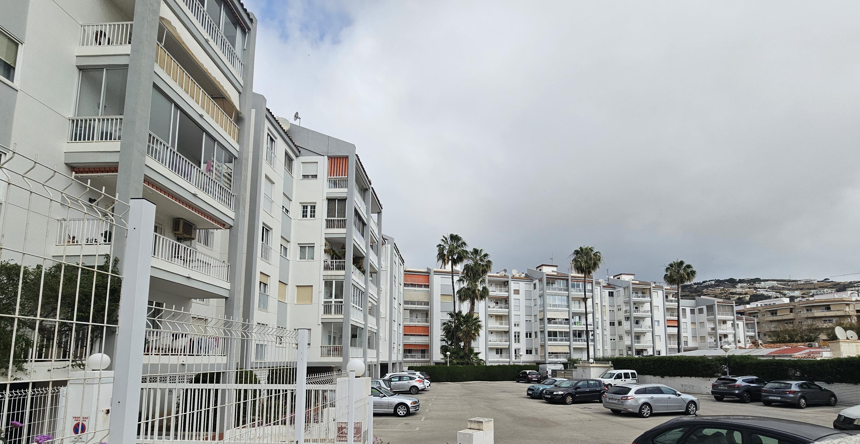Vista exterior del edificio residencial con balcones privados, zona de aparcamiento comunitario vallada y palmeras bajo un cielo despejado en Jávea.