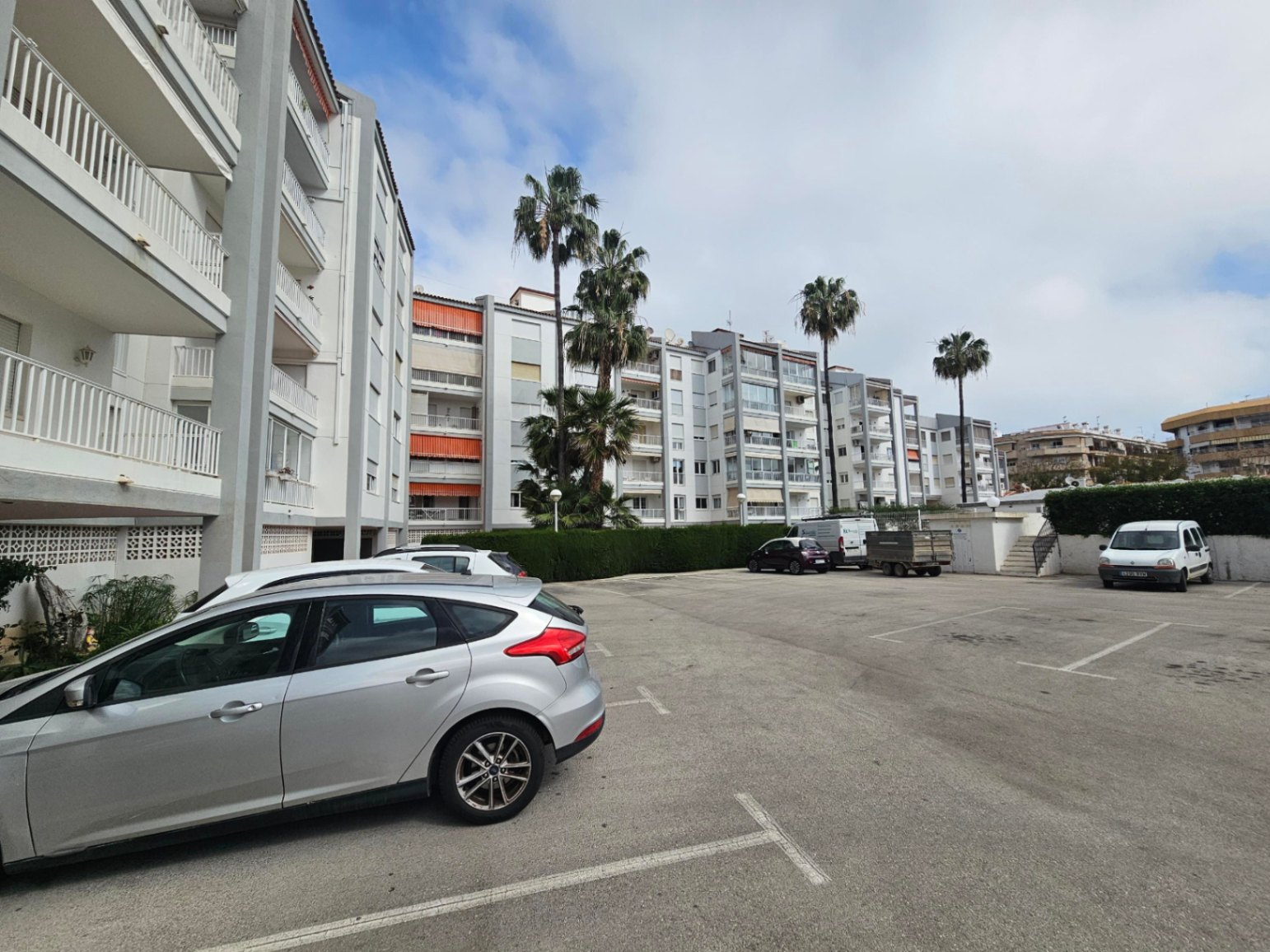 Zona de aparcamiento comunitario exterior con plazas delimitadas, rodeada de setos cuidados y palmeras frente a la fachada del edificio residencial.