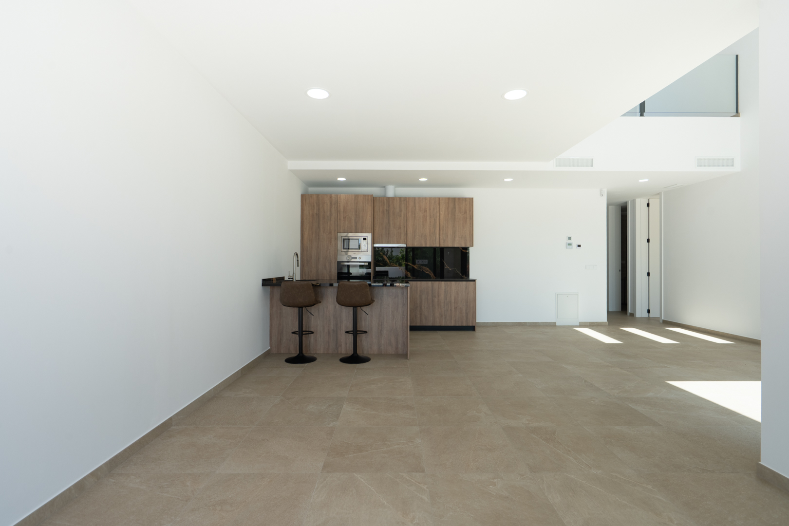 Cocina de diseño abierta con armarios de madera, encimera oscura y barra con taburetes, integrada en un amplio salón con suelos porcelánicos y techos de doble altura.