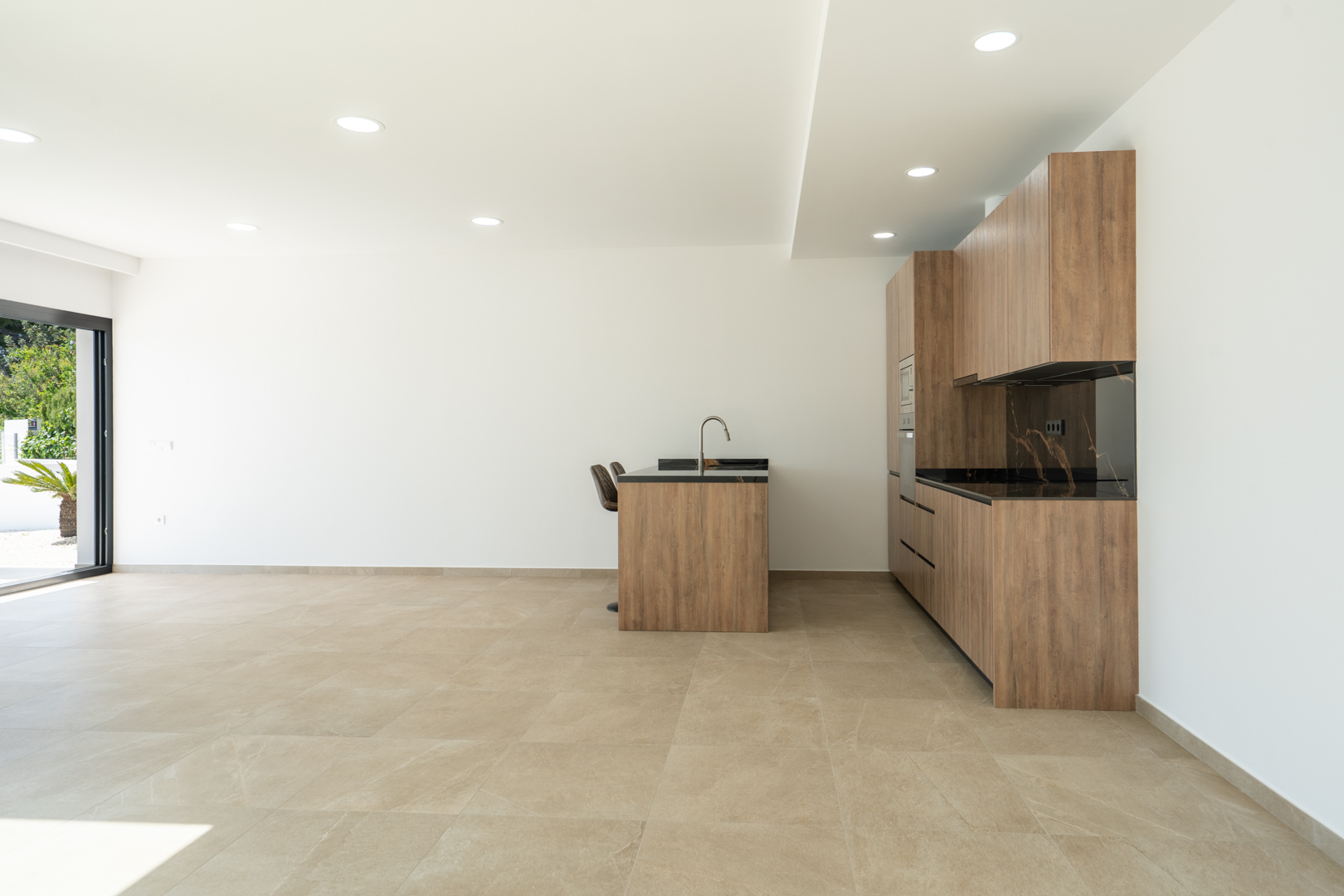 Cocina de diseño con mobiliario en acabado madera, encimera oscura veteada e isla central, integrada en un espacio diáfano con suelos porcelánicos y luz empotrada.
