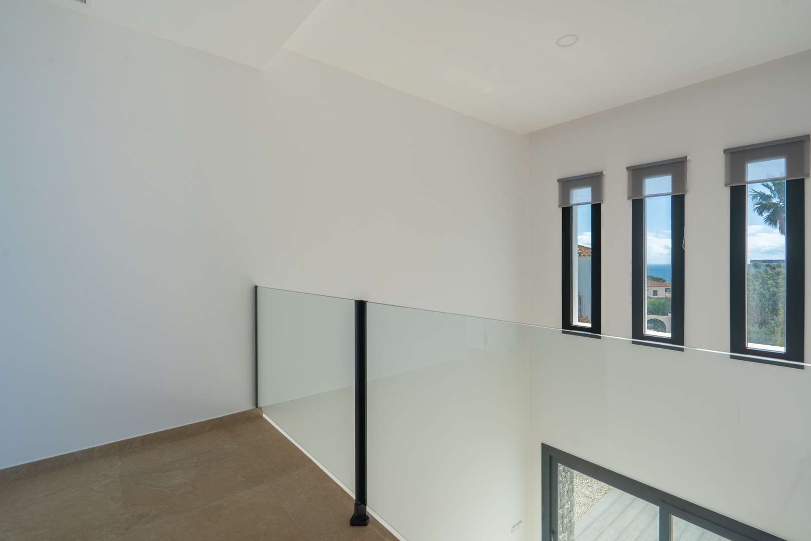 Distribuidor de la planta superior con barandilla de cristal y vistas al mar a través de tres ventanales verticales. Suelo porcelánico y techos con iluminación empotrada.