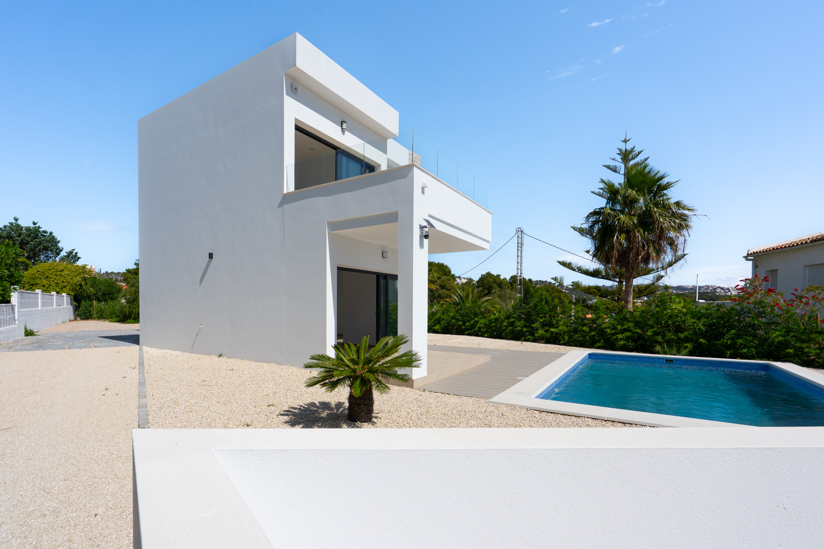Exterior de la villa moderna con fachada blanca, piscina rectangular y amplias terrazas con barandillas de cristal sobre una parcela llana con jardín de grava.