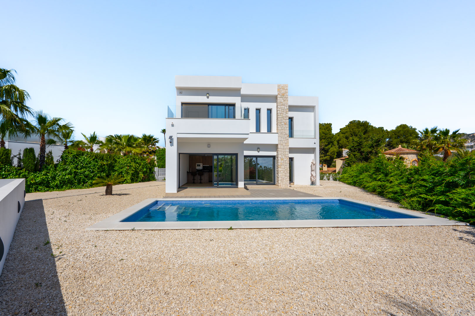 Fachada moderna de dos niveles con detalles en piedra natural, piscina rectangular rodeada de grava y amplios ventanales que conectan el interior con el exterior.