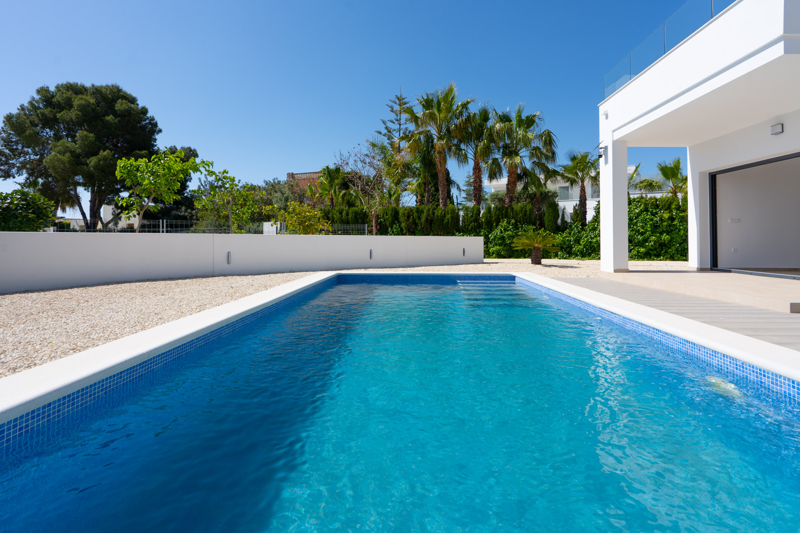 Piscina rectangular de gresite azul rodeada de grava blanca y muros perimetrales modernos con iluminación integrada, junto a vegetación mediterránea.