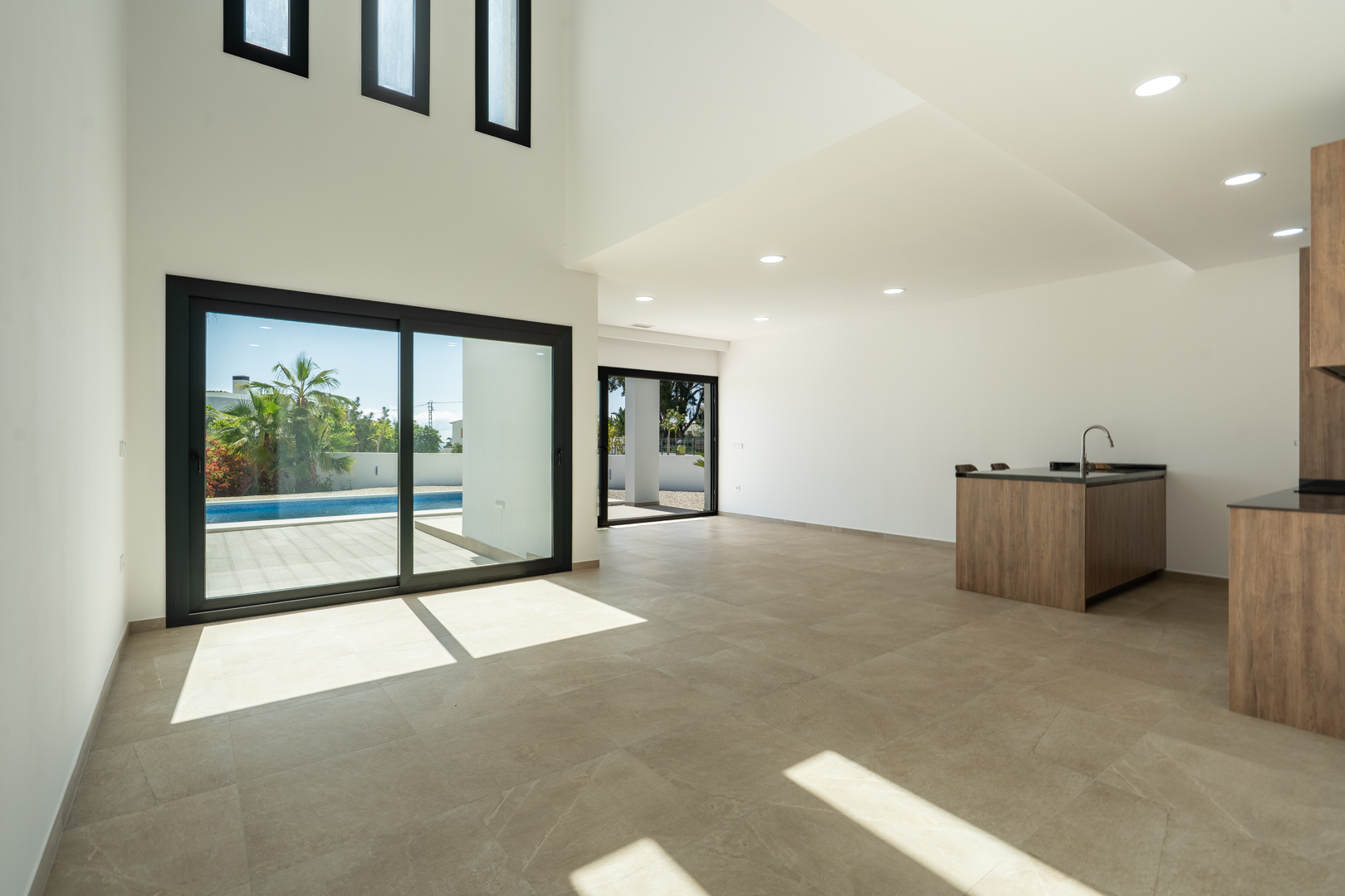 Salón-comedor de doble altura con cocina abierta, suelos porcelánicos y grandes ventanales que conectan directamente con la terraza y la piscina.