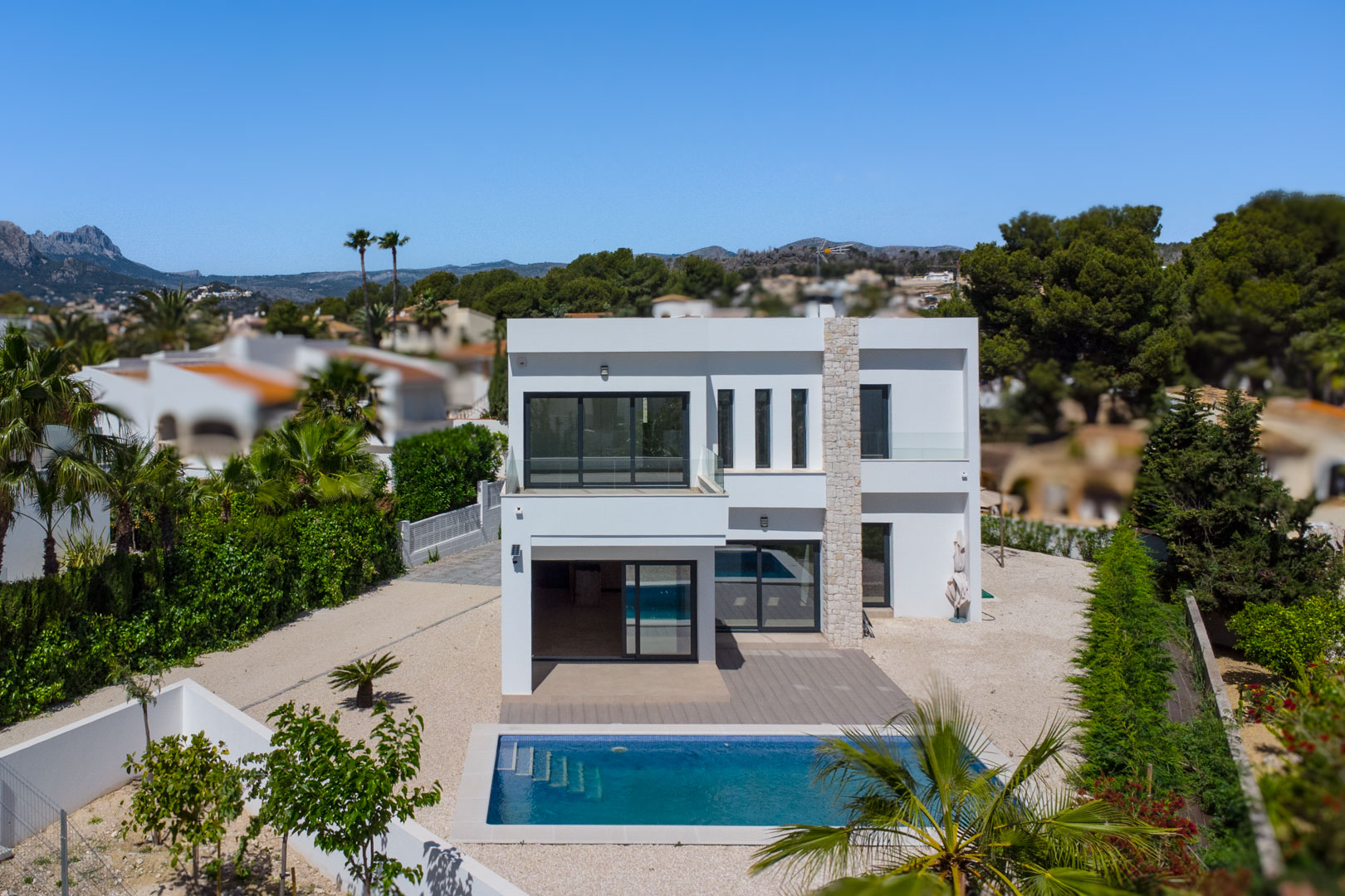 Vista exterior de la villa moderna con fachada blanca, pilar de piedra natural, piscina rectangular y amplias terrazas con barandillas de cristal.