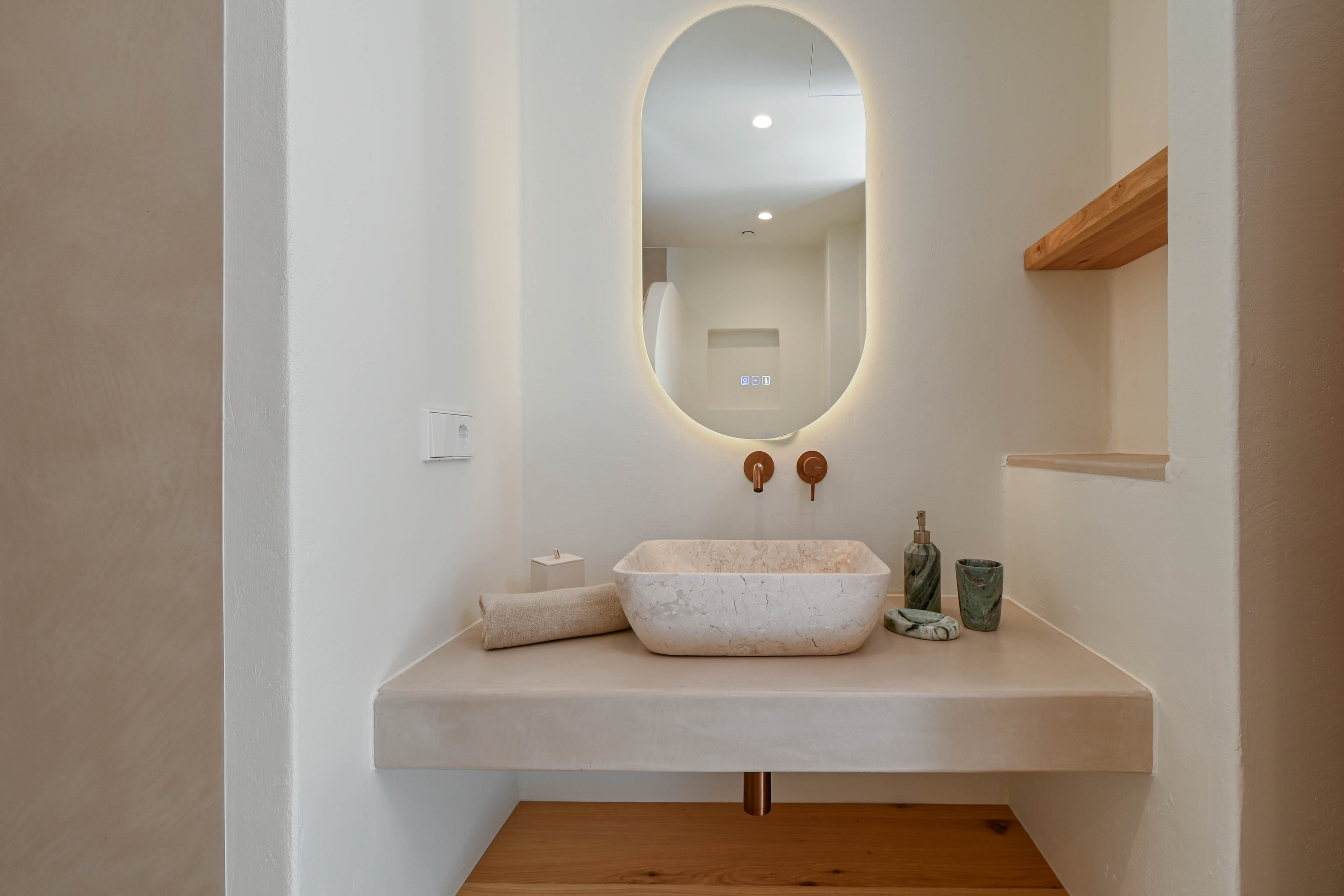 Baño de diseño con lavabo de piedra natural sobre encimera de microcemento, grifería empotrada en cobre y espejo ovalado retroiluminado con detalles en madera.