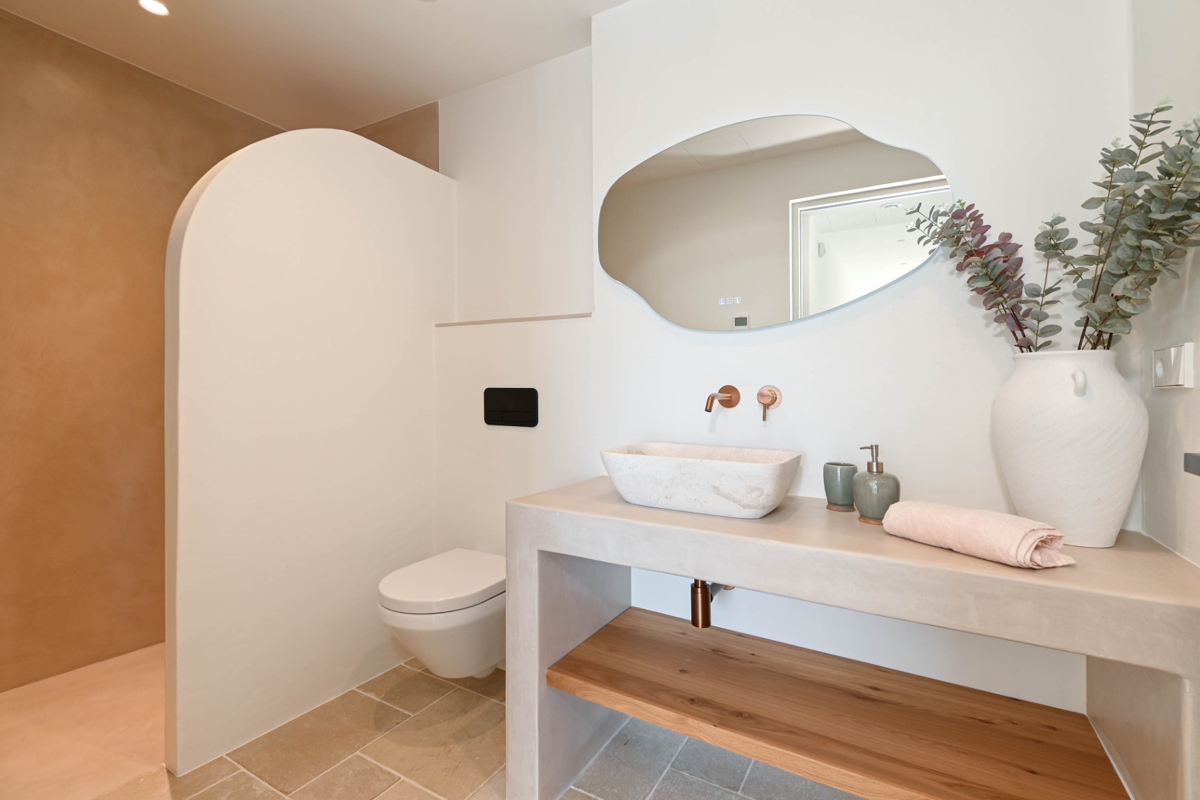 Baño moderno con lavabo de piedra sobre encimera de microcemento, grifería empotrada en cobre, espejo orgánico y zona de ducha con tabique curvo y acabados naturales.