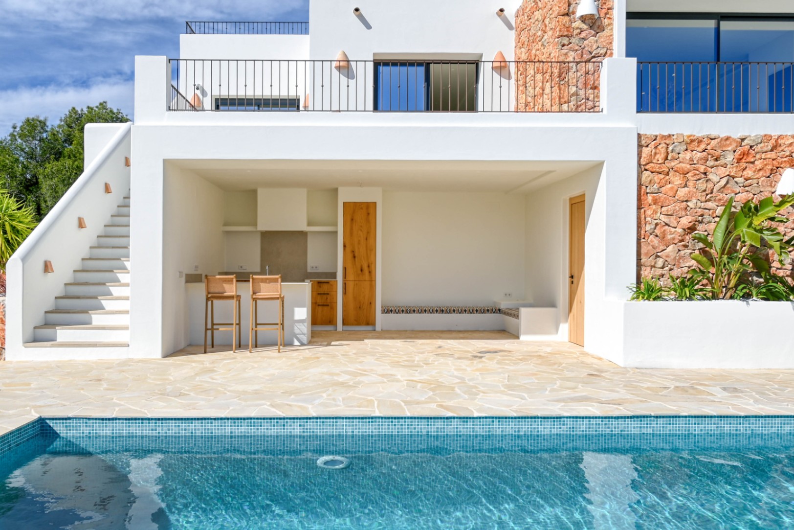 Cocina exterior equipada y zona de estar junto a la piscina, con acabados en piedra natural, madera y suelos de losa irregular bajo una fachada blanca moderna.