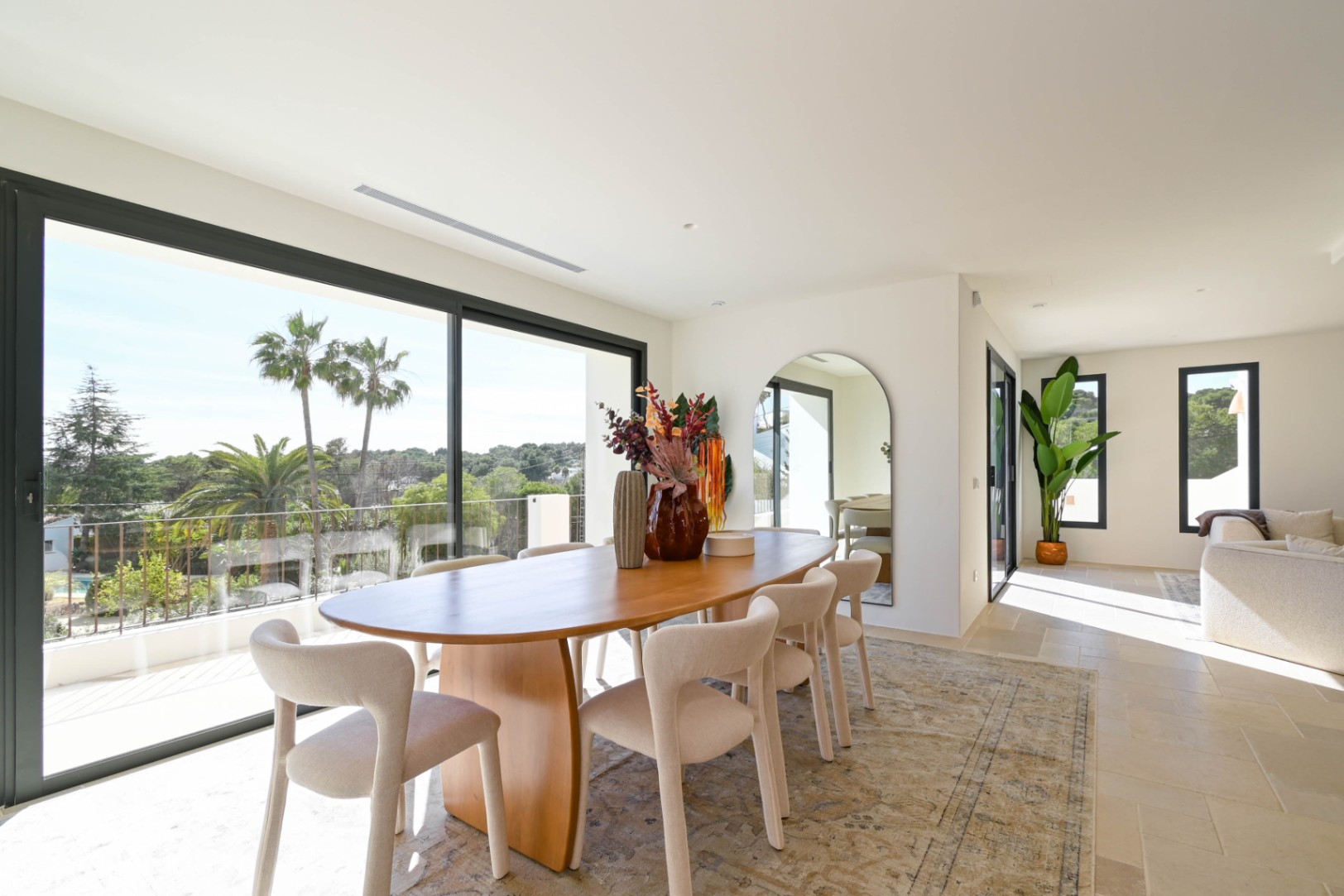 Comedor luminoso con mesa ovalada de madera y amplios ventanales correderos que ofrecen vistas despejadas a las palmeras y al entorno natural de Moraira.