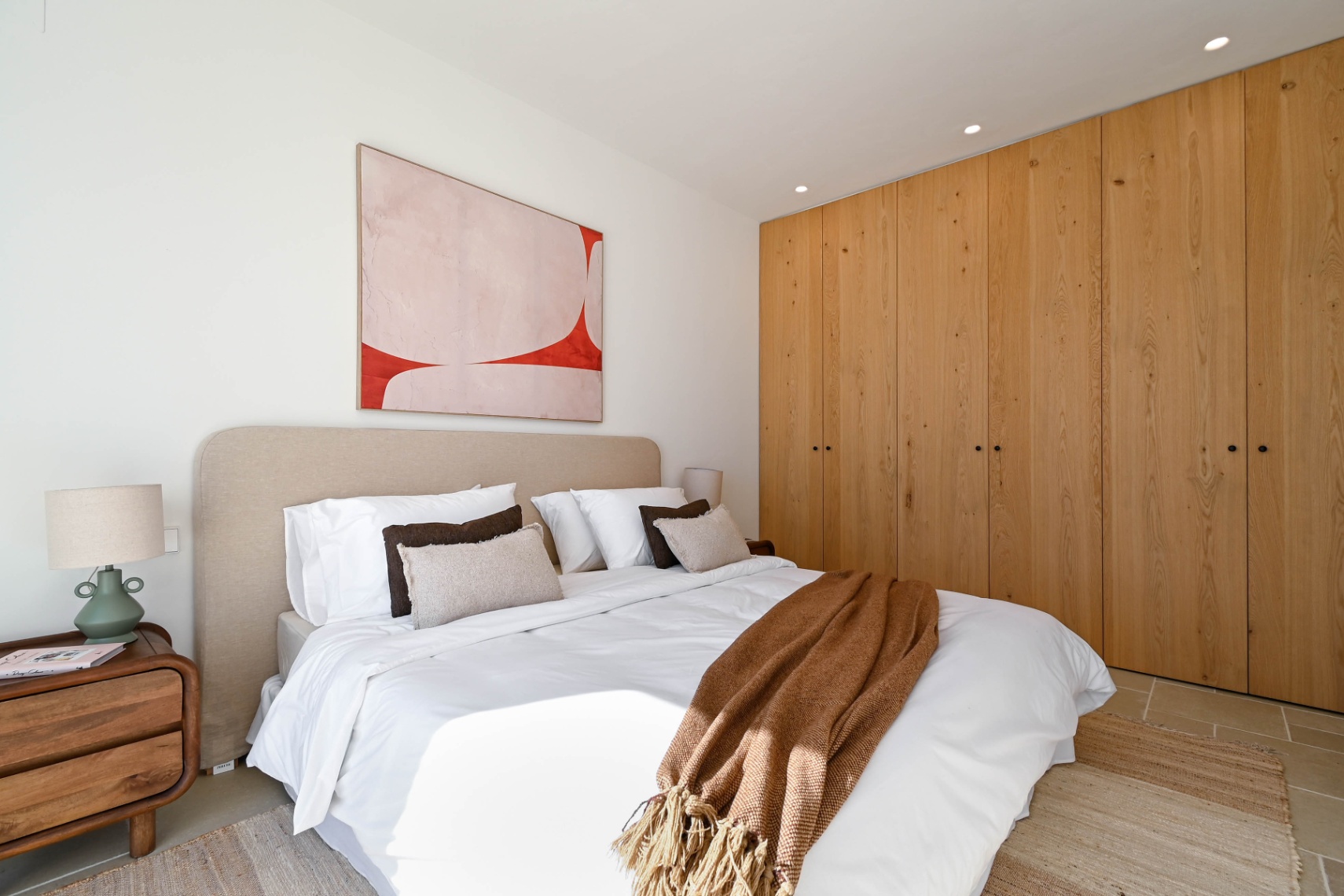 Dormitorio con armarios empotrados de madera natural de suelo a techo, cabecero tapizado en beige e iluminación integrada. El diseño incluye suelos cerámicos y una decoración minimalista.